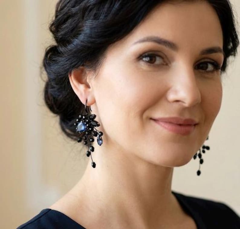May include: A pair of dangling earrings featuring black metal wire, small black beads, and a central dark blue teardrop-shaped crystal. The earrings have a cluster design with multiple thin strands extending downward, creating a layered appearance.