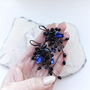 May include: A pair of dangle earrings featuring clusters of small black beads and faceted royal blue teardrop crystals. The components are attached to thin black wire stems that extend outward, creating a textured, organic shape with a dark finish.