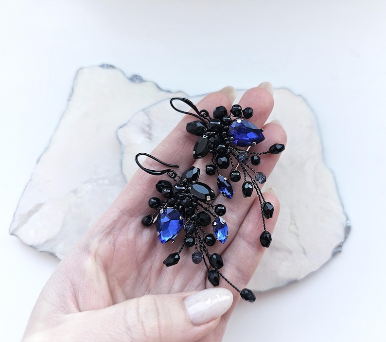 May include: A pair of dangle earrings featuring clusters of small black beads and faceted royal blue teardrop crystals. The components are attached to thin black wire stems that extend outward, creating a textured, organic shape with a dark finish.
