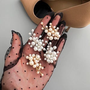 May include: Four pearl cluster brooches with gold-tone wire accents are displayed on a black, polka-dotted glove. The brooches feature various sizes of white and cream-colored pearls. A pair of beige high heels is in the background.