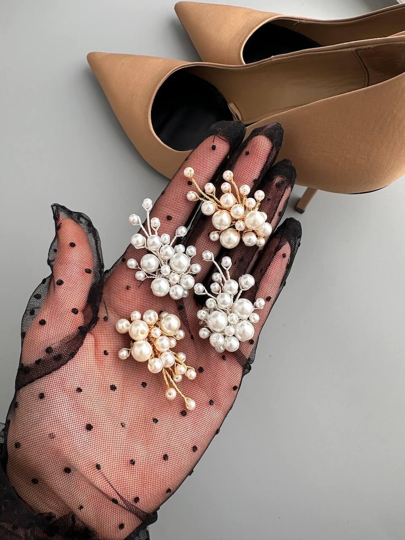 May include: Four pearl cluster brooches with gold-tone wire accents are displayed on a black, polka-dotted glove. The brooches feature various sizes of white and cream-colored pearls. A pair of beige high heels is in the background.