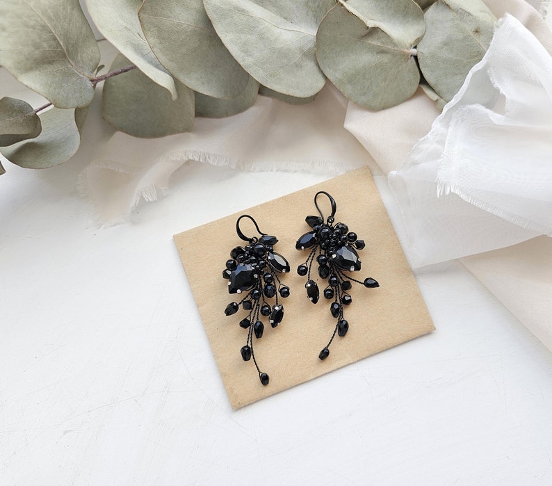 May include: A pair of black dangle earrings featuring clusters of faceted black beads and crystals attached to thin black wire stems. The earrings have black hook-style ear wires and are arranged in a cascading, branch-like formation.