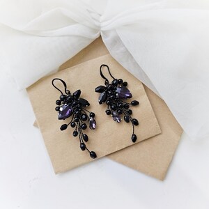 May include: A pair of dangle earrings featuring clusters of black beads and faceted purple crystals. The earrings are constructed with thin black wire and include black metal ear hooks for suspension, creating a cascading, branch-like appearance.