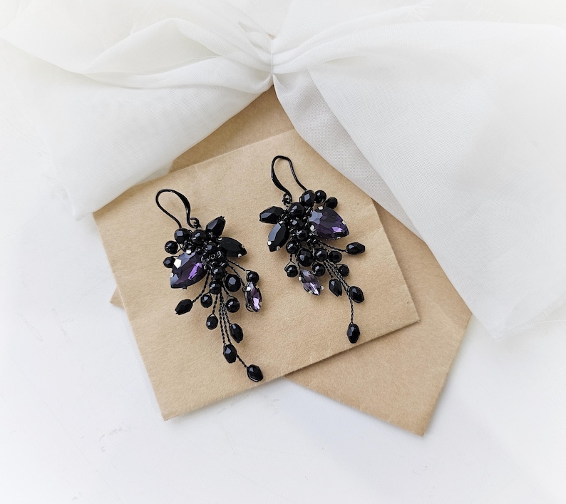 May include: A pair of dangle earrings featuring clusters of black beads and faceted purple crystals. The earrings are constructed with thin black wire and include black metal ear hooks for suspension, creating a cascading, branch-like appearance.