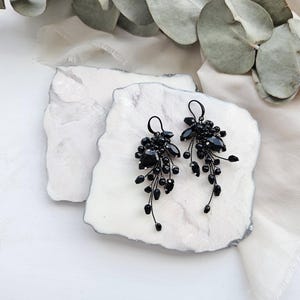 May include: A pair of dangling earrings featuring clusters of black faceted beads and teardrop-shaped stones attached to thin black wire stems. The earrings have black metal ear hooks and a cascading design that tapers toward the bottom of each piece.