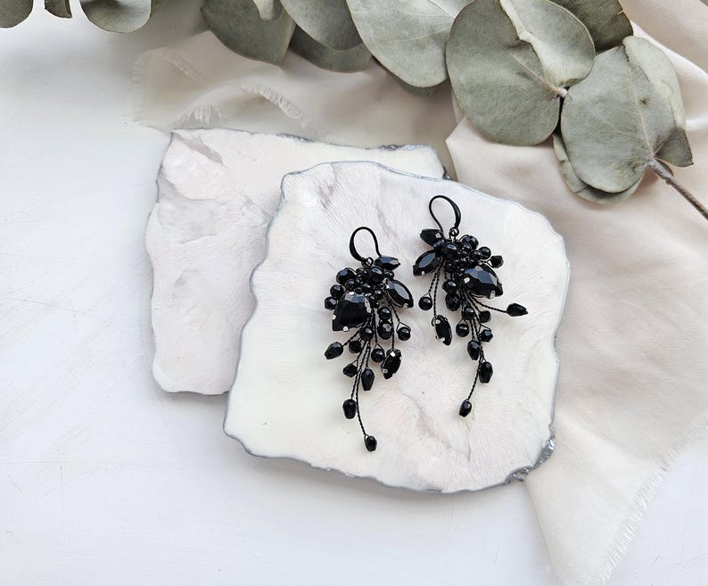 May include: A pair of dangling earrings featuring clusters of black faceted beads and teardrop-shaped stones attached to thin black wire stems. The earrings have black metal ear hooks and a cascading design that tapers toward the bottom of each piece.