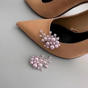 May include: Tan pointed-toe heels with a cluster of light pink faux pearls and silver wire accents. One cluster is attached to the shoe, and another is placed nearby. The shoes have a classic, elegant design.