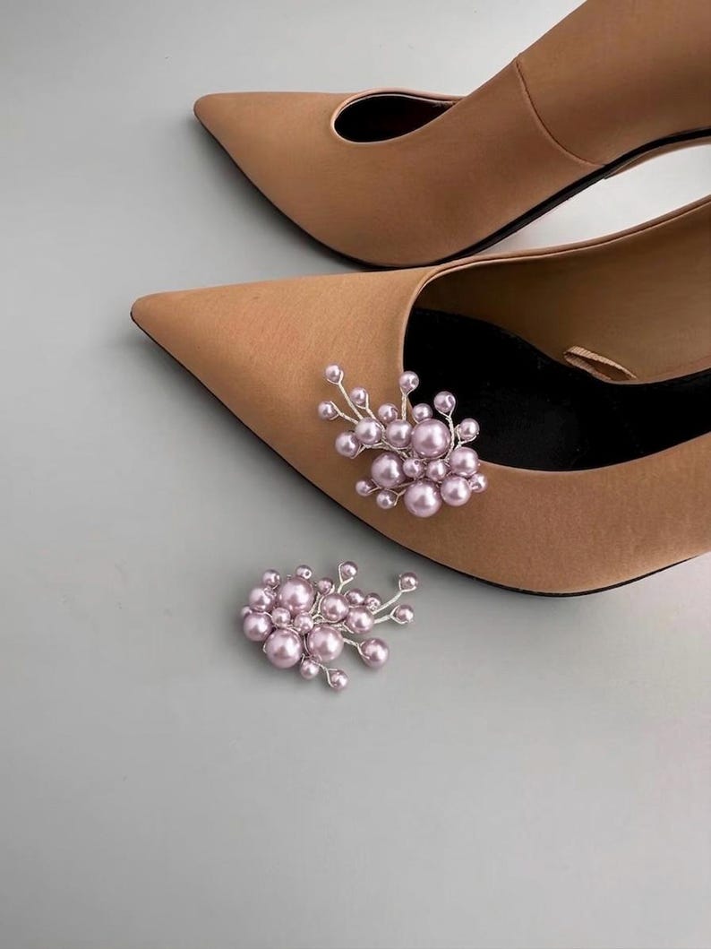 May include: Tan pointed-toe heels with a cluster of light pink faux pearls and silver wire accents. One cluster is attached to the shoe, and another is placed nearby. The shoes have a classic, elegant design.