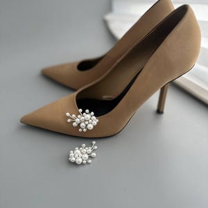 May include: A pair of tan high-heeled pumps with pointed toes, featuring a small cluster of white pearls attached to the front of each shoe. The heels are slender and the shoes are set against a gray background.