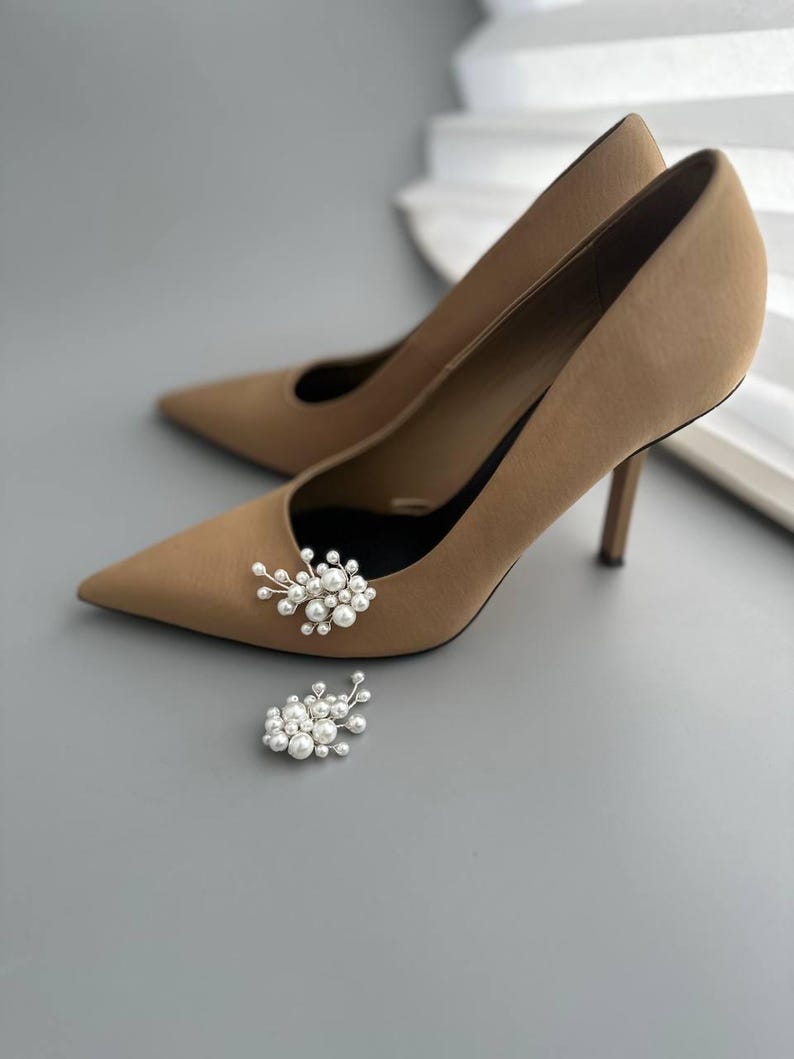 May include: A pair of tan high-heeled pumps with pointed toes, featuring a small cluster of white pearls attached to the front of each shoe. The heels are slender and the shoes are set against a gray background.