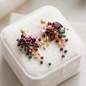 May include: A pair of gold-tone earrings featuring clusters of deep red, emerald green, and pale pink crystals. The design includes teardrop-shaped stones, small round beads, and delicate gold wire branches that extend outward in a spray.