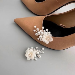 May include: Tan pointed-toe heels with floral shoe clips. The shoe clips feature white fabric flowers and faux pearls. The heels are a neutral color, suitable for various outfits and occasions. The background is a light gray.