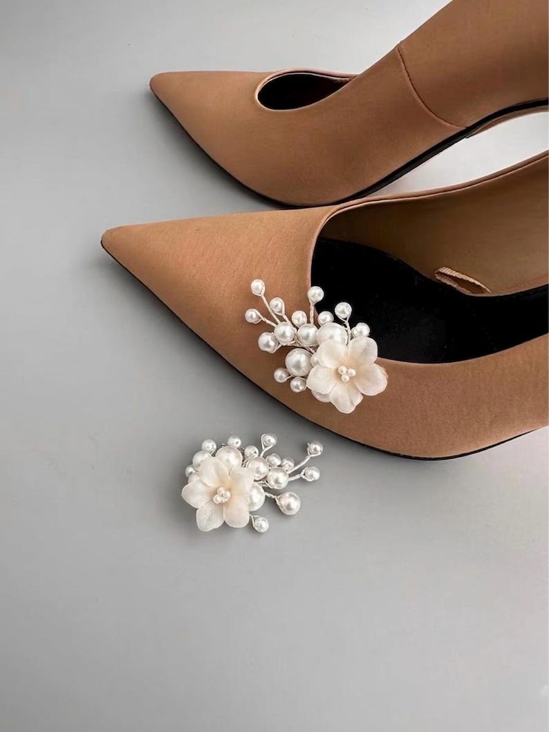 May include: Tan pointed-toe heels with floral shoe clips. The shoe clips feature white fabric flowers and faux pearls. The heels are a neutral color, suitable for various outfits and occasions. The background is a light gray.