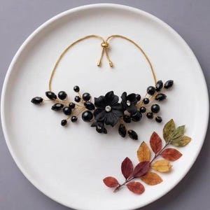 May include: A gold-toned adjustable necklace featuring black floral accents, black beads, and small clear crystals. The floral centrepiece is composed of two black flowers with central crystals, surrounded by black beads and gold wire branches.