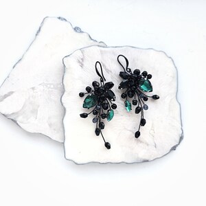 May include: A pair of dangling earrings featuring black wire branches adorned with small black beads and faceted emerald green teardrop crystals. The earrings have black metal hooks and a cascading, botanical-inspired design with multiple dark accents.