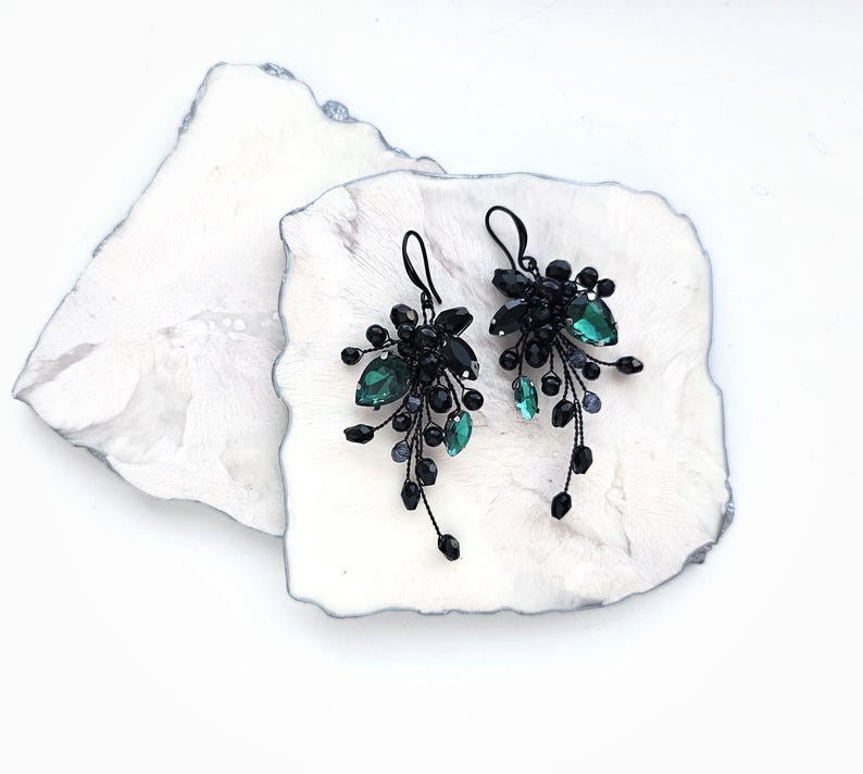 May include: A pair of dangling earrings featuring black wire branches adorned with small black beads and faceted emerald green teardrop crystals. The earrings have black metal hooks and a cascading, botanical-inspired design with multiple dark accents.