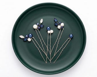 May include: A set of four silver-toned hair pins featuring decorative clusters of navy blue pearls, white pearls, and clear marquise-cut crystals. Each pin has a U-shaped wire base with a wavy design, arranged in a fan pattern on a dark green plate.