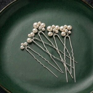 May include: Six silver hair pins with clusters of faux pearls. The pins are arranged on a dark green ceramic plate. The pearls vary in size, creating a delicate and elegant look. Ideal for weddings or special occasions.