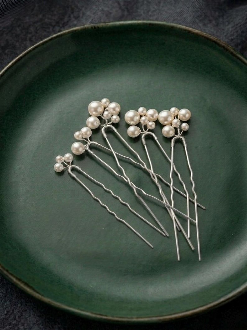 May include: Six silver hair pins with clusters of faux pearls. The pins are arranged on a dark green ceramic plate. The pearls vary in size, creating a delicate and elegant look. Ideal for weddings or special occasions.