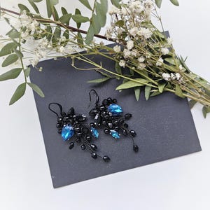 May include: A pair of dangle earrings featuring clusters of small black beads and faceted black stones, accented with prominent teardrop-shaped blue crystals. The earrings are suspended from black metal hooks and have a delicate, branching design.