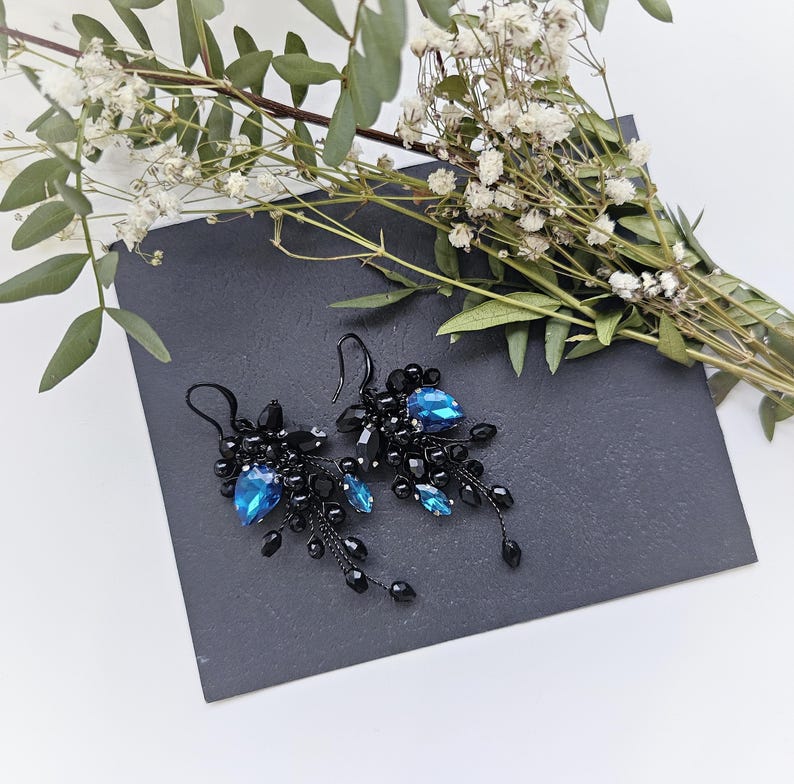 May include: A pair of dangle earrings featuring clusters of small black beads and faceted black stones, accented with prominent teardrop-shaped blue crystals. The earrings are suspended from black metal hooks and have a delicate, branching design.