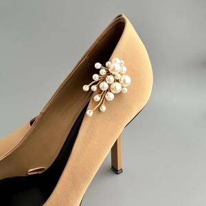 May include: A pair of tan high-heeled shoes with a cluster of faux pearls and gold-colored stems attached to the side. The shoes have a pointed toe and a stiletto heel. The background is a solid gray.