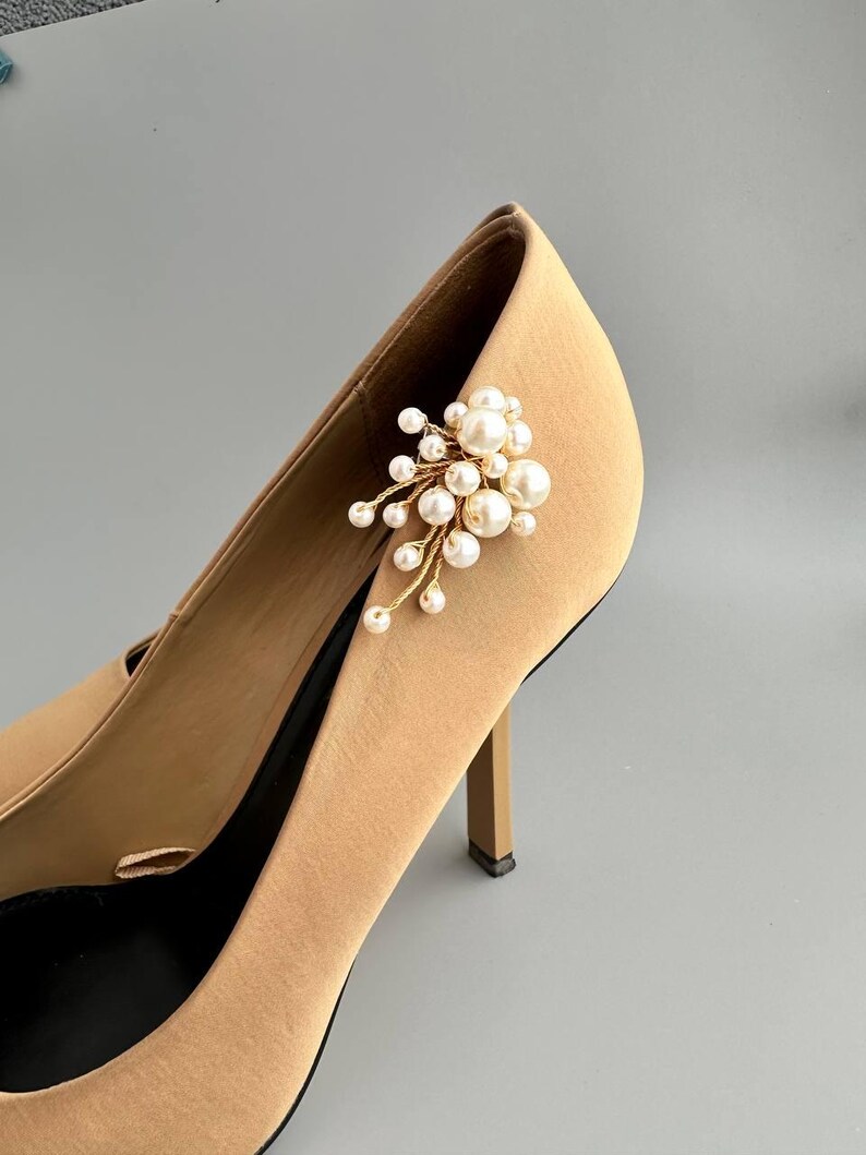 May include: A pair of tan high-heeled shoes with a cluster of faux pearls and gold-colored stems attached to the side. The shoes have a pointed toe and a stiletto heel. The background is a solid gray.