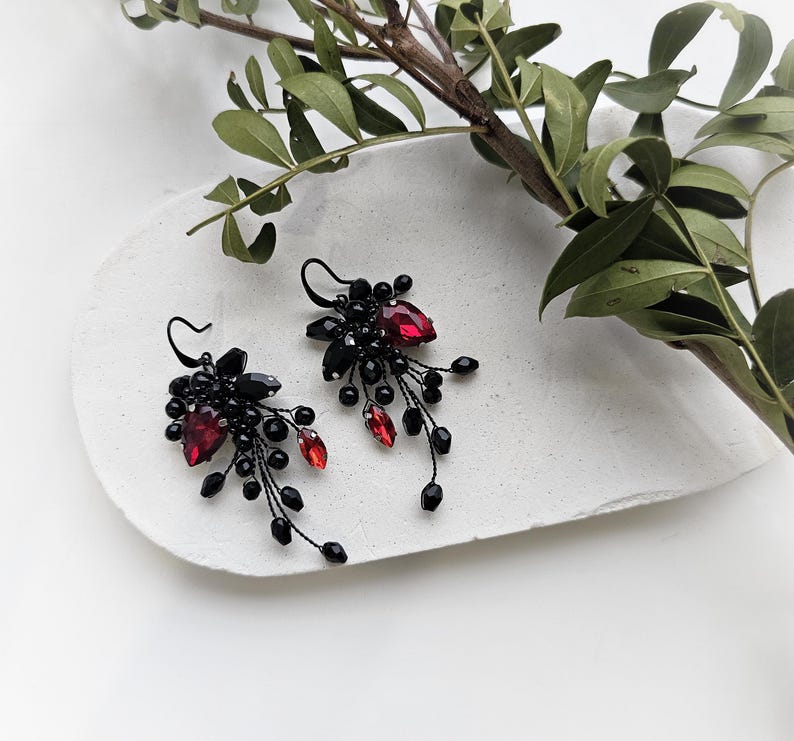 May include: A pair of dangle earrings featuring black wire branches adorned with small black beads and faceted red teardrop crystals. The earrings have black metal hooks and a cluster design that extends downward with delicate wire stems and beads.