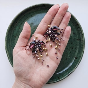 May include: A pair of cluster earrings featuring large, teardrop-shaped purple crystals, smaller round purple beads, and metallic gold accents. The earrings are constructed with thin gold-toned wire, creating a delicate, branching arrangement of beads and crystals that extend outward from the central teardrop stones.