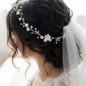 May include: A silver-toned hair vine featuring delicate white floral accents and small round white pearls. The flexible wire accessory is woven through dark hair and is paired with a sheer white tulle veil attached to the back of the hairstyle.
