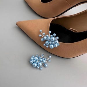 May include: A pair of tan pointed-toe heels with a light blue pearl embellishment. A matching pearl embellishment is placed separately. The shoes are on a light gray surface.