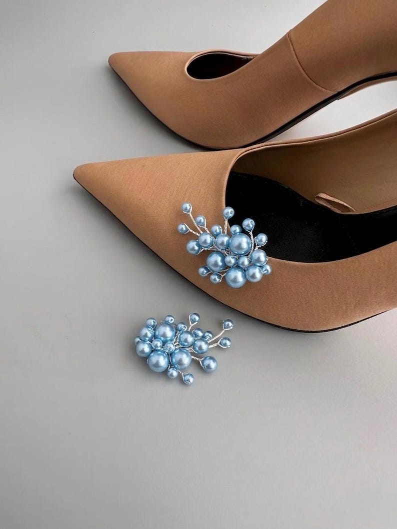 May include: A pair of tan pointed-toe heels with a light blue pearl embellishment. A matching pearl embellishment is placed separately. The shoes are on a light gray surface.