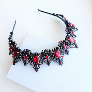 Puede incluir: Una corona de cuentas negras y rojas con grandes piedras preciosas rojas ovaladas. La corona presenta intrincados detalles de filigrana y está colocada sobre una diadema negra. Las piedras preciosas son de color rojo intenso y las cuentas son negras.