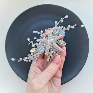 Puede incluir: Un peine de pelo plateado con un diseño de estrella de mar, adornado con cristales azules y blancos. El peine se sostiene en una mano sobre un fondo negro.