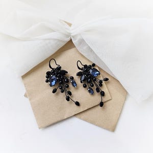 May include: A pair of dangle earrings featuring dark blue faceted crystals and small black beads arranged in a cluster. The earrings are attached to black metal hooks and have a cascading design with delicate wire branches extending downward.