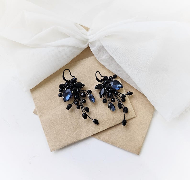 May include: A pair of dangle earrings featuring dark blue faceted crystals and small black beads arranged in a cluster. The earrings are attached to black metal hooks and have a cascading design with delicate wire branches extending downward.