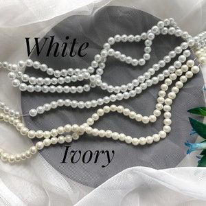 May include: Strands of white and ivory faux pearls are displayed against a gray background with the words "White" and "Ivory" in black script. The pearls are strung together, showcasing their round shape and glossy finish.