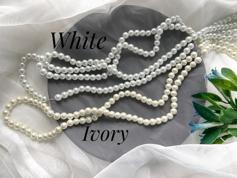 May include: Strands of white and ivory faux pearls are displayed against a gray background with the words "White" and "Ivory" in black script. The pearls are strung together, showcasing their round shape and glossy finish.