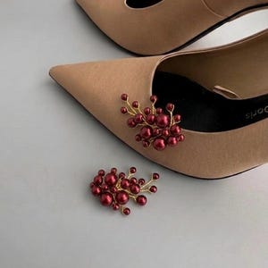 May include: A pair of beige pointed-toe pumps featuring a decorative cluster of deep red spherical beads attached to the front of one shoe, with a matching loose cluster of red beads placed separately on the surface next to the footwear.