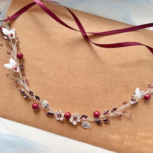 May include: A flexible hair vine featuring silver-toned wire, deep red pearls, clear crystal accents, and metallic silver leaf charms. The piece is adorned with small dark red faceted beads and includes long burgundy satin ribbons at both ends.