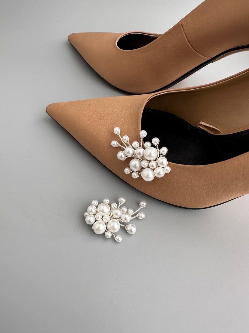 May include: A pair of tan pointed-toe heels with a pearl and silver embellishment. A matching pearl and silver embellishment is next to the shoe. The shoes are on a gray background.