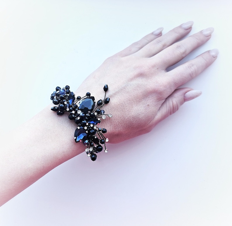 May include: A bracelet featuring dark blue and black faceted crystals, small clear rhinestones, and black beads arranged in a cluster on a thin wire frame. The design includes various shapes of stones, including teardrop and round cuts, creating a dense, textured appearance across the wrist area.