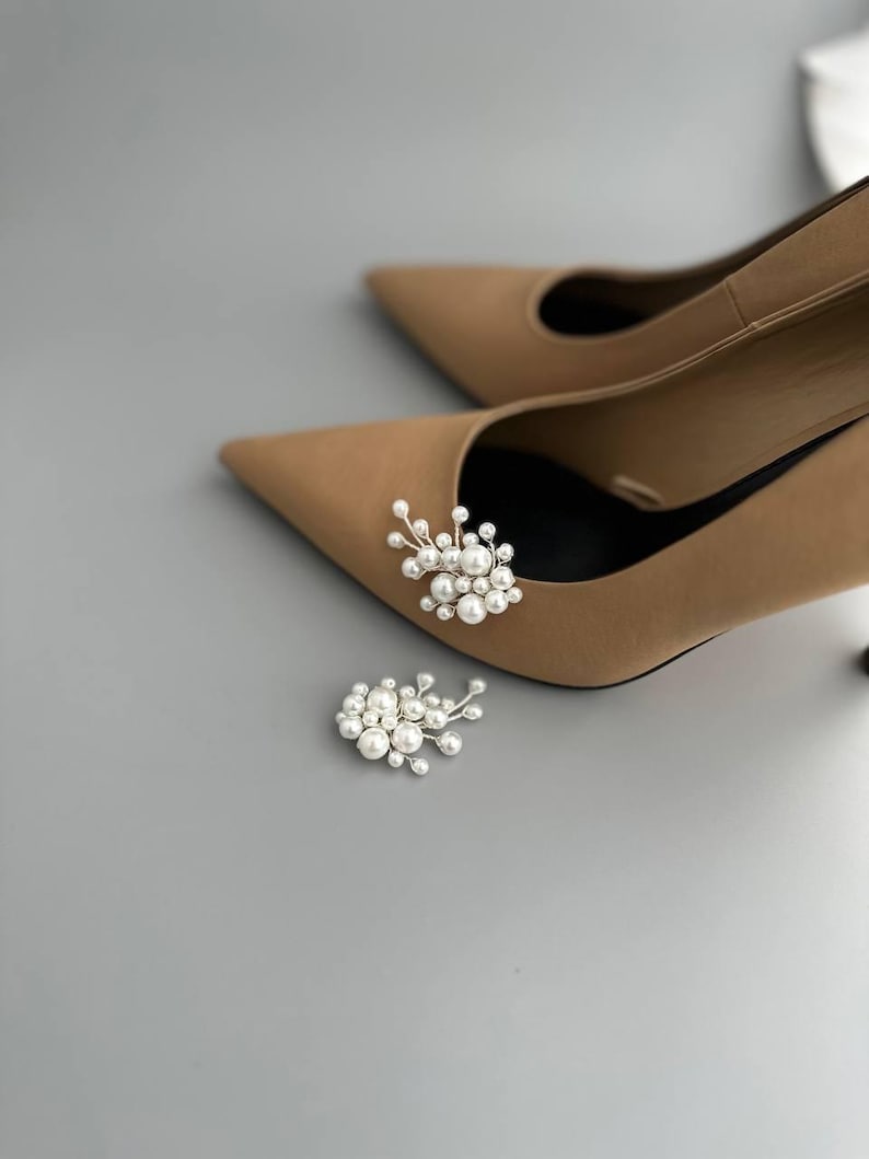 May include: Tan pointed-toe high heels with pearl embellishments. One shoe has a pearl cluster attached, and another cluster rests on the surface. The heels are on a gray background.