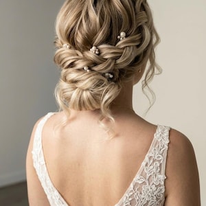 May include: A blonde updo hairstyle with a braided crown and a low bun, adorned with small pearl hairpins. The woman is wearing a white lace dress with a deep V-neck.