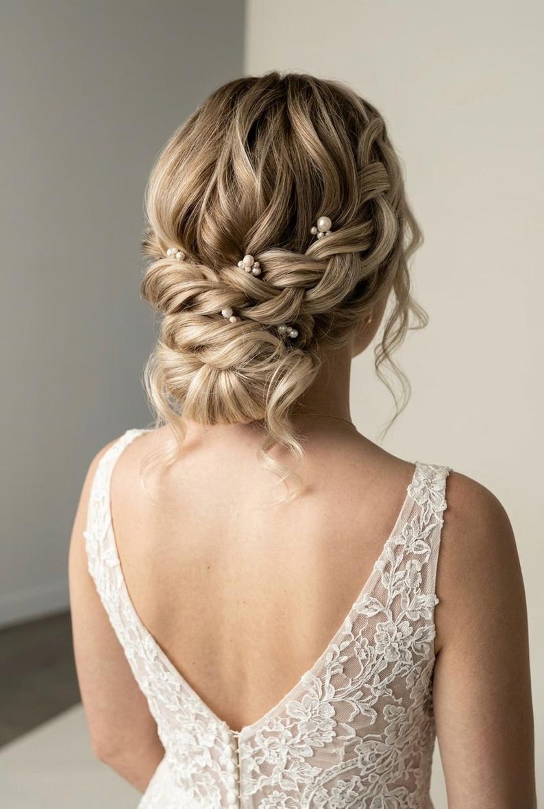 May include: A blonde updo hairstyle with a braided crown and a low bun, adorned with small pearl hairpins. The woman is wearing a white lace dress with a deep V-neck.