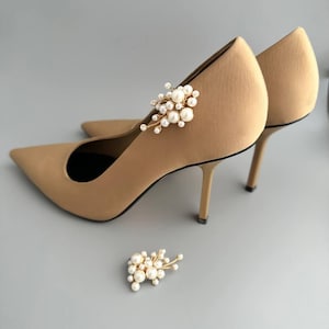 May include: Tan-colored pointed-toe high heels with a delicate pearl and gold embellishment. The heels have a stiletto design and are set against a gray background. A matching pearl and gold embellishment is placed near the heels.