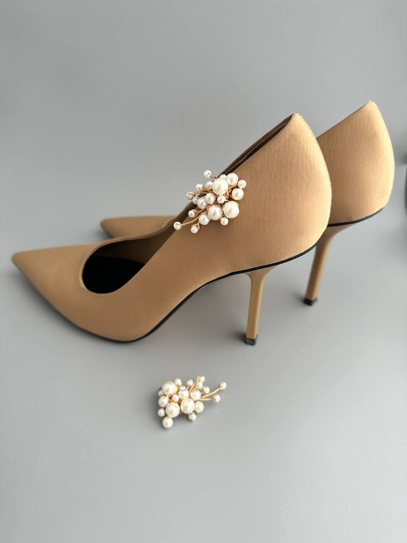 May include: Tan-colored pointed-toe high heels with a delicate pearl and gold embellishment. The heels have a stiletto design and are set against a gray background. A matching pearl and gold embellishment is placed near the heels.