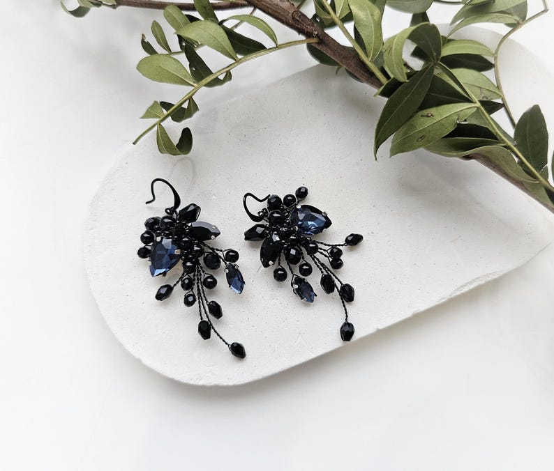 May include: A pair of dangle earrings featuring dark black wire, small black beads, and faceted deep blue crystal stones arranged in a cluster design. The earrings have a hook-style closure and a delicate, branching structure with multiple dangling elements.