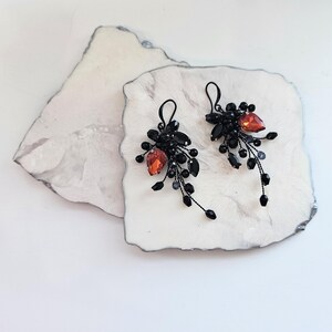 May include: A pair of dangle earrings featuring black wire branches adorned with small black beads and a single prominent teardrop-shaped orange crystal on each piece. The earrings have black metal hooks for attachment and a cluster design.