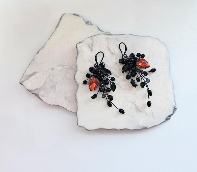 May include: A pair of dangle earrings featuring black wire branches adorned with small black beads and a single prominent teardrop-shaped orange crystal on each piece. The earrings have black metal hooks for attachment and a cluster design.